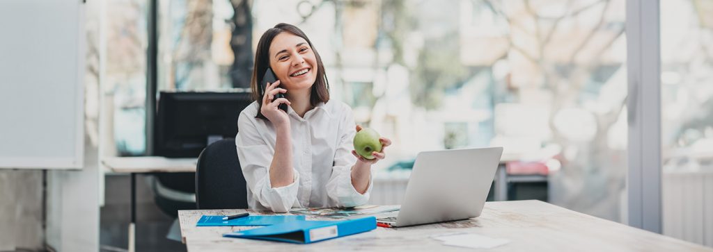 Eine junge Frau telefoniert in einem hellen Büro mit einem grünen Apfel in der Hand