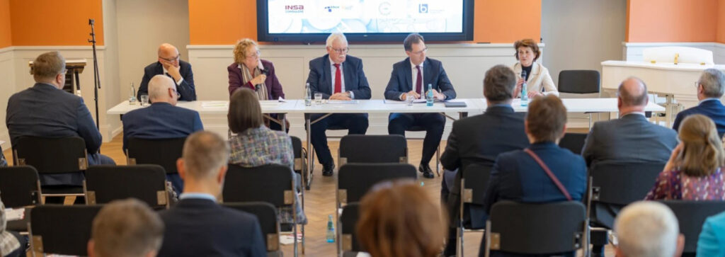 fünf Personen sitzen an einem langen Tisch und sprechen zu den Gästen