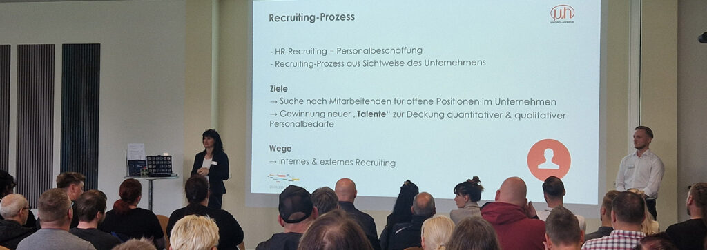 Teilnehmerinnen und Teilnehmer sitzen in einem großen Raum und verfolgen eine Präsentation.